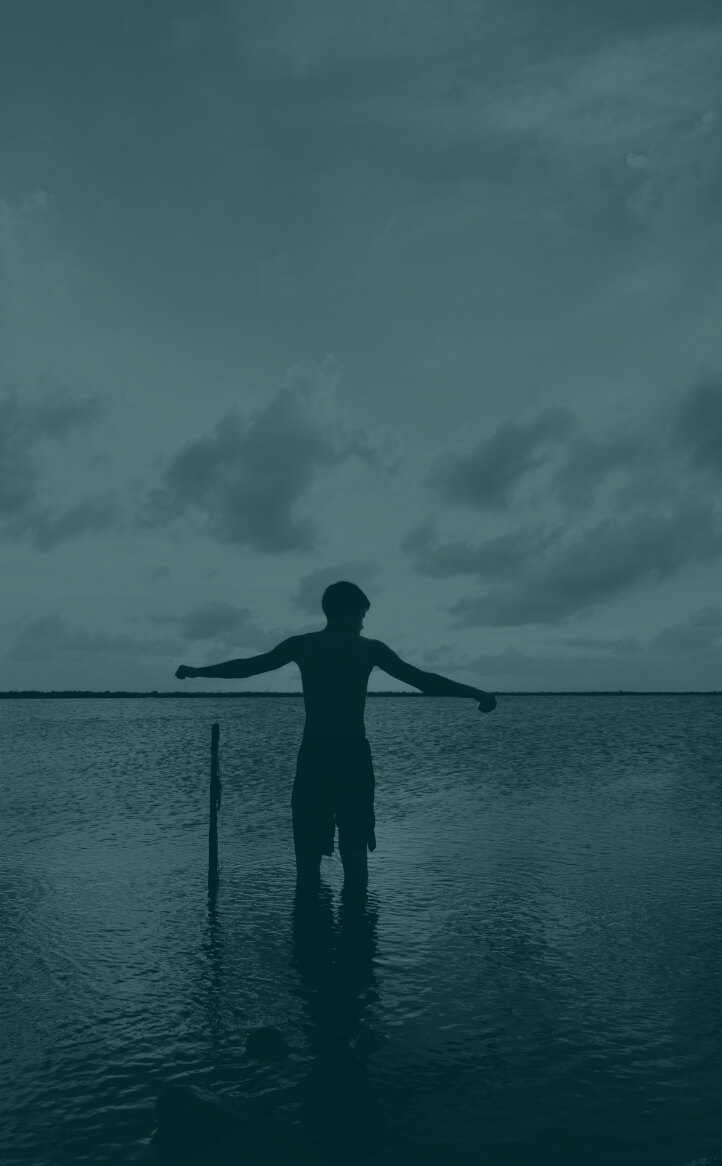 A person stands in shallow water with arms outstretched, gazing at a cloudy sky over Louisiana chemical industry heartland.