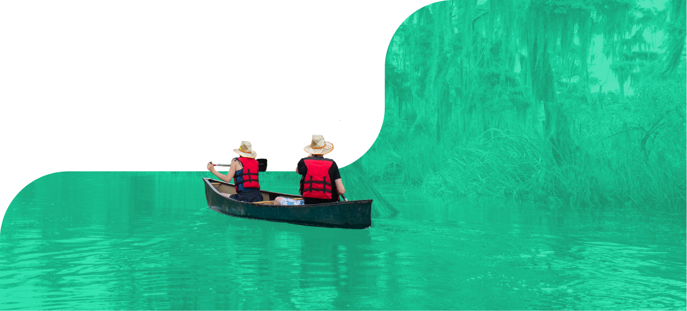 Two people wearing life jackets and hats paddle a canoe along Louisiana's industrial corridor, surrounded by dense, green-tinted foliage.