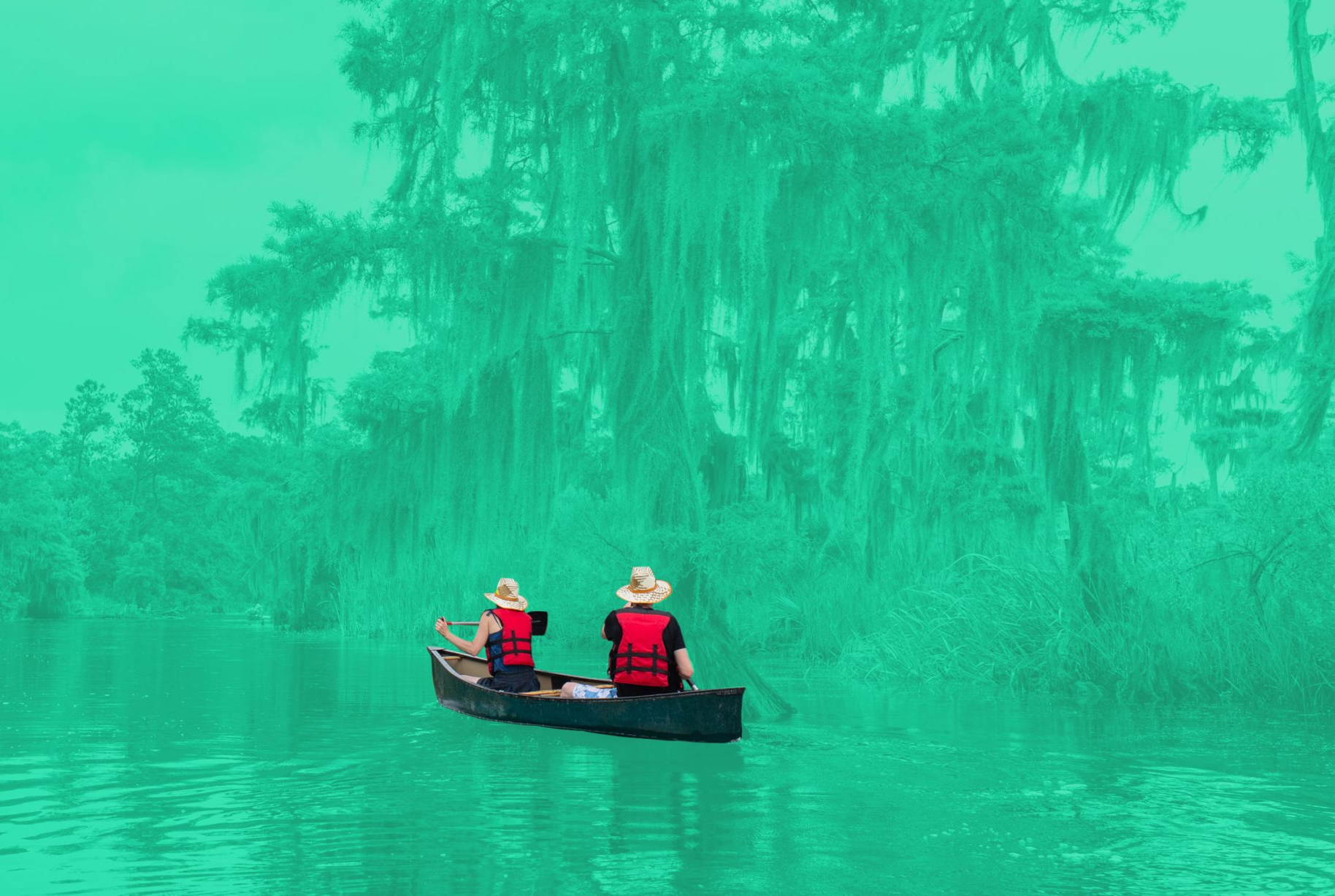 Two people in red life jackets paddle a canoe on a calm river, surrounded by trees and moss near the Louisiana industrial corridor.